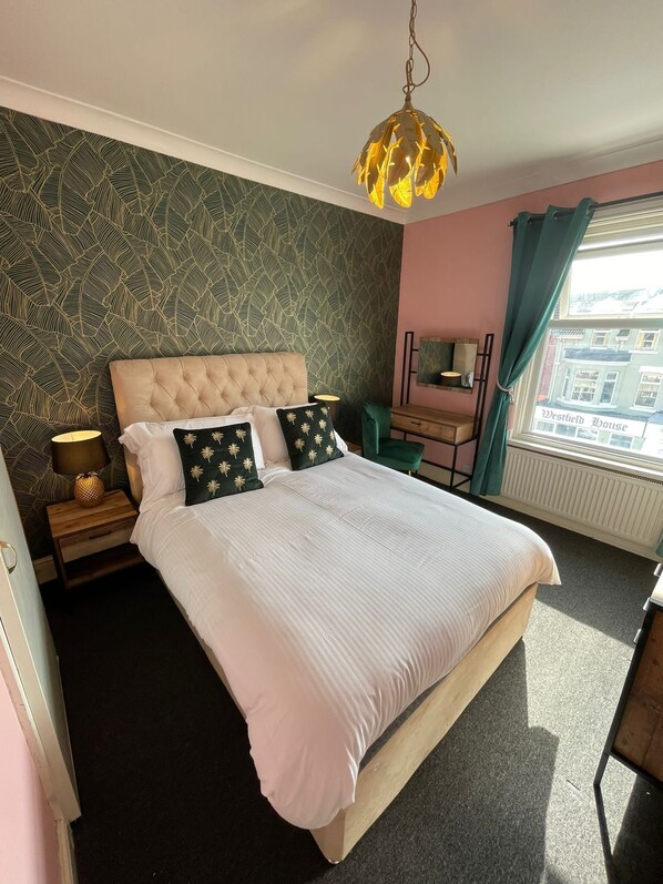A bedroom with a tufted headboard, a large bed with white bedding, decorative pillows, a bedside lamp, a dresser, and a view of the city through a window.