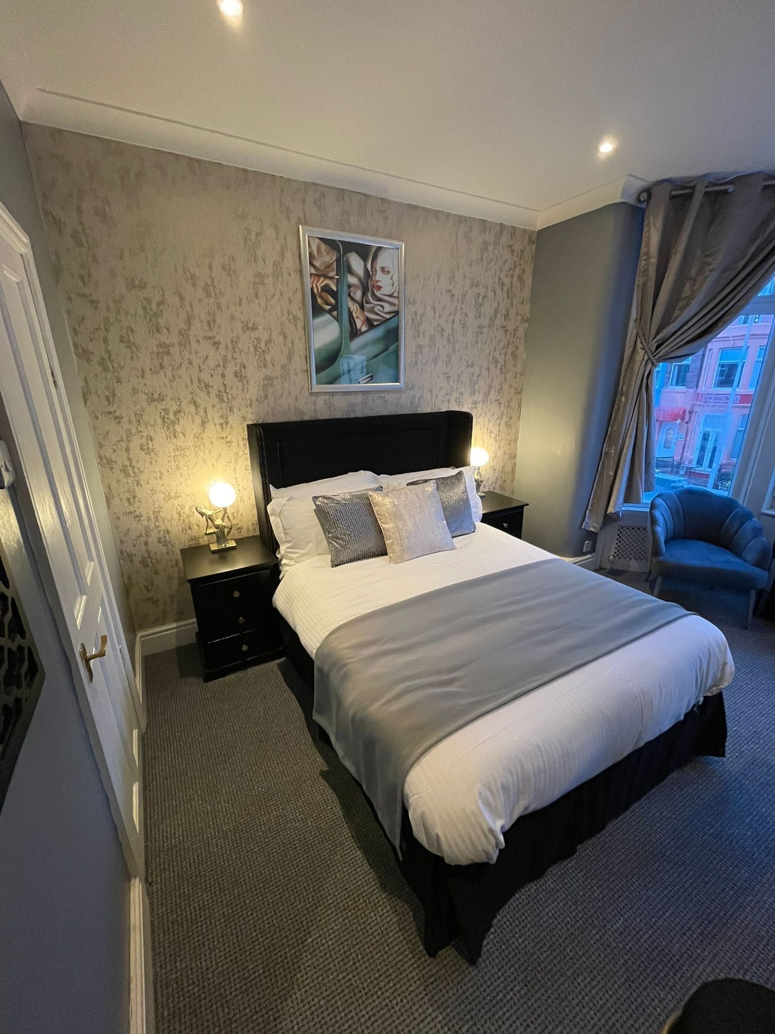 Une chambre d’hôtel avec un grand lit, une table de chevet avec une lampe, un tableau au mur et une vue sur les bâtiments par la fenêtre.