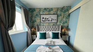 A neatly made bed with a blue quilt, white pillows, and a white towel arranged in a bow on the bed. There is a framed picture on the wall above the bed. - McGraths Blackpool (Blackpool)