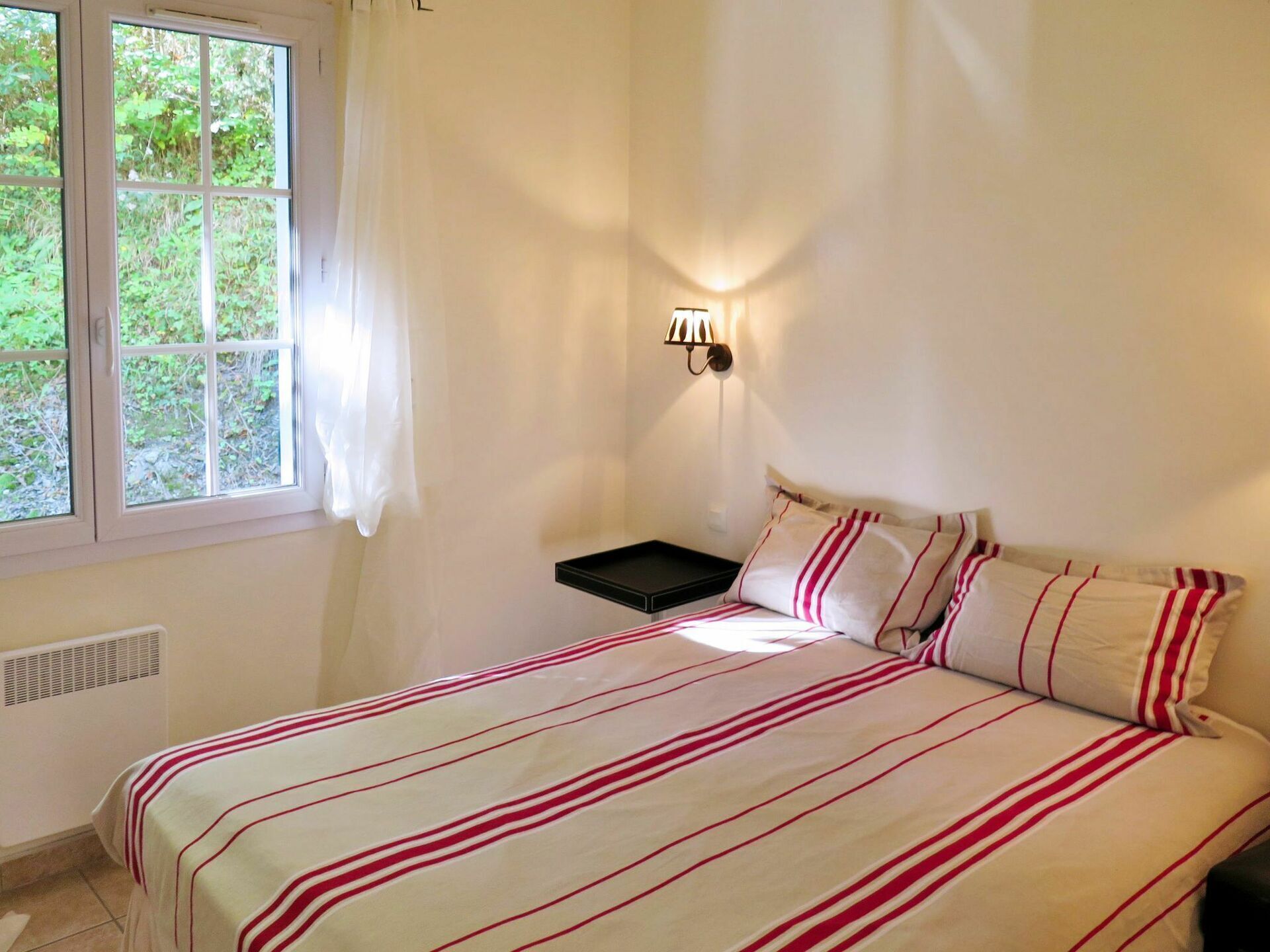 A bedroom with a bed, a nightstand, a wall-mounted lamp, and a window with curtains.