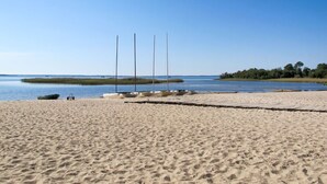 Ein sandiger Strand mit ruhigem Gewässer, einigen Booten und einer fernen Baumlinie.