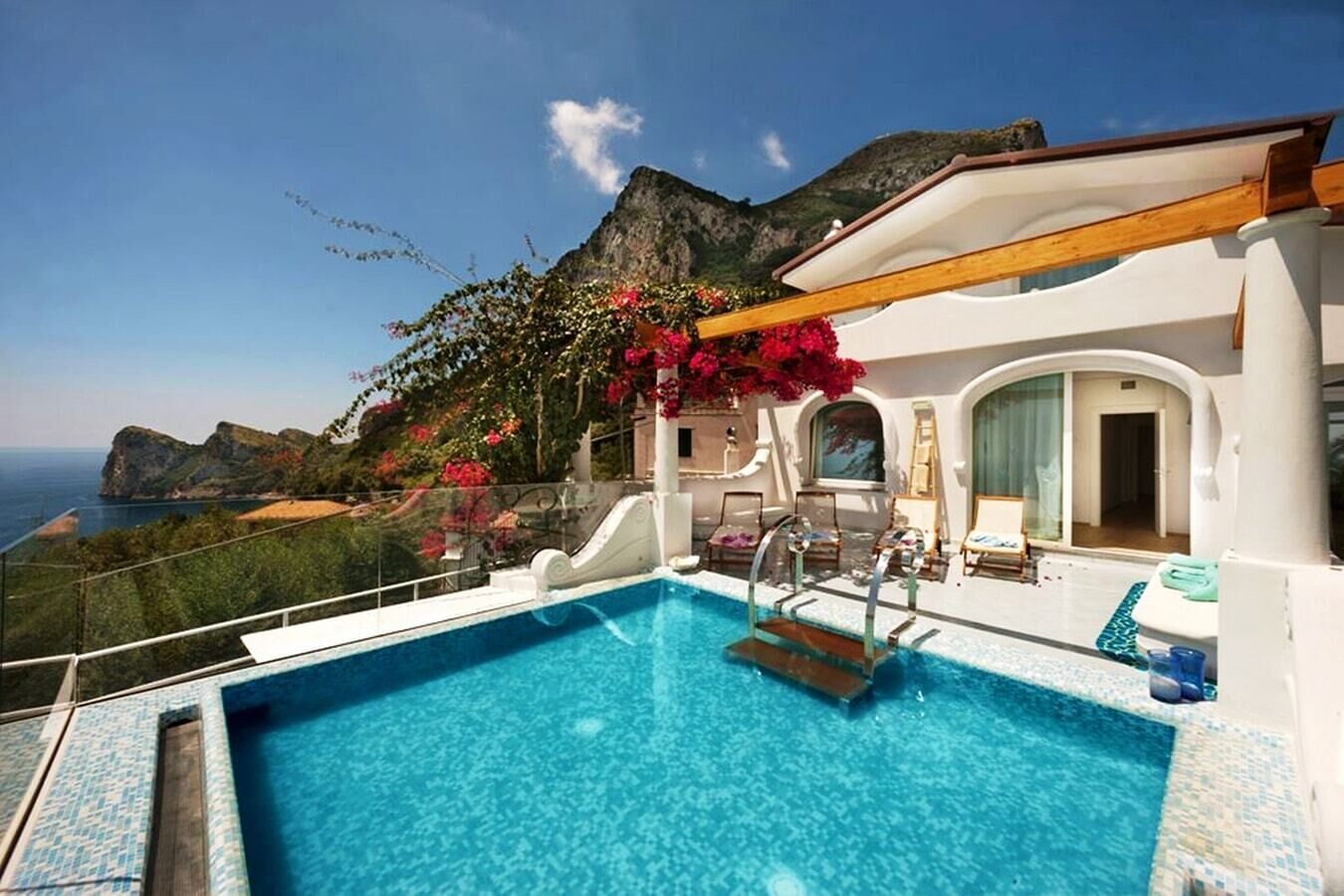 A pool with a view of the sea and mountains, a covered area with seating, and a building with a white facade.