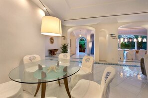 Zona pranzo con tavolo in vetro, sedie bianche e una decorazione a stella marina. Sono presenti archi e vista sull'esterno.