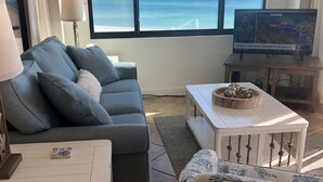 Un salon avec un canapé gris, une table basse blanche et un fauteuil à motifs. Un téléviseur est posé sur un meuble en bois, et une fenêtre offre une vue sur l’océan.