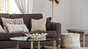 Een moderne woonkamer met een donkerbruine bank, gestreepte kussens, een glazen salontafel en een geweven voetenbankje.