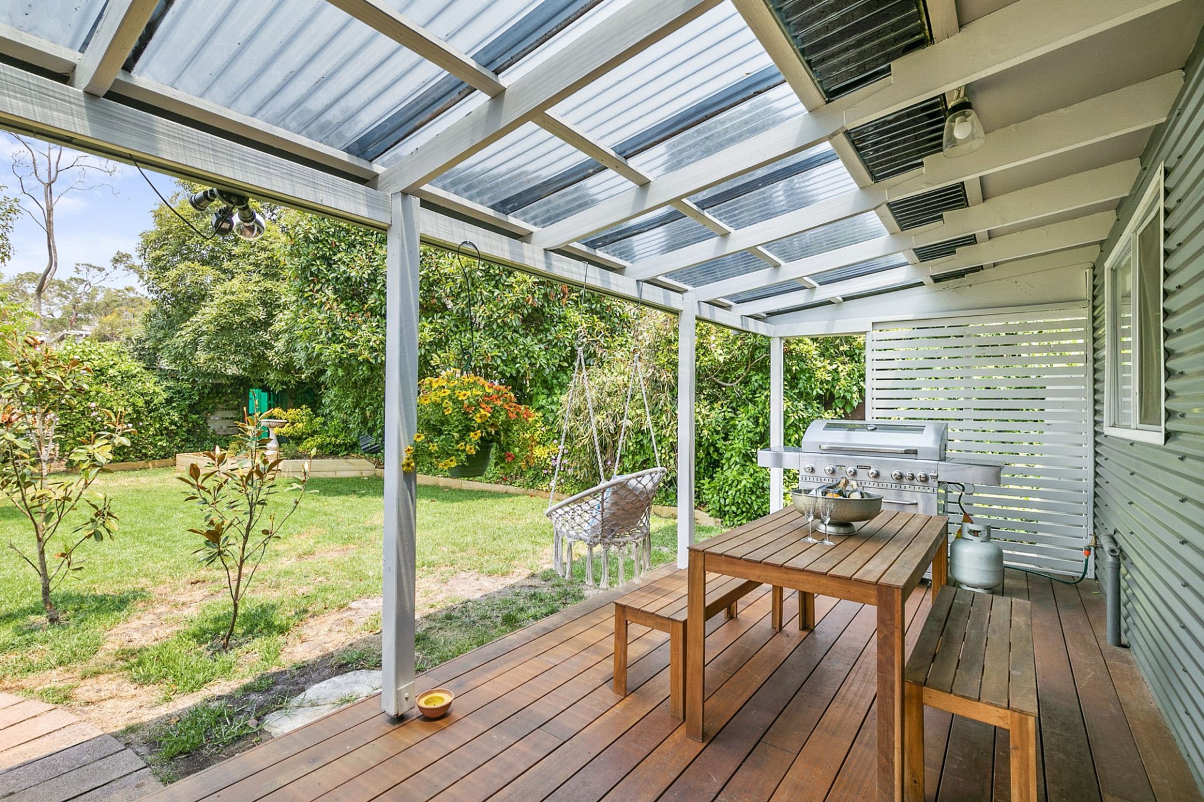 A covered outdoor patio with a wooden table and chairs, a grill, and a view of a garden.