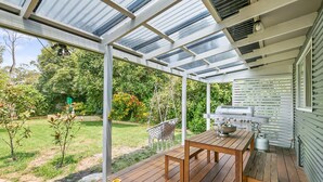 Eine überdachte Terrasse im Freien mit einem Holztisch und -stühlen, einem Grill und Blick auf einen Garten.