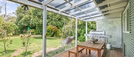 Un patio techado al aire libre con mesa y sillas de madera, una parrilla y vistas a un jardín.