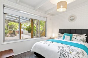 A bedroom with a large window, a bed with floral bedding, a bedside table with a lamp, and a hanging light fixture. - The Beach House Anglesea (Anglesea)