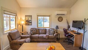 Ein Wohnzimmer mit einer Couch, einem Holztisch mit Obstkorb, einem Schreibtisch mit Computer und zwei Fenstern mit Jalousien.