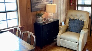 A wooden interior with a dining table, chairs, a cushioned armchair, a lamp, a painting, and a fire extinguisher. - Once a Part of the Beloved Shady Oak Lodge - Cabin 3 w/ Boat Ramp (Many)
