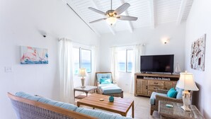 Un vaste espace de vie agrémenté d’un ventilateur de plafond, d’un téléviseur, de meubles en osier et offrant une vue sur la mer grâce à de grandes fenêtres.
