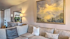 Een woonkamer met een beige bank, een groot schilderij van een bos en een houten kast met decoratieve objecten.