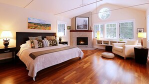 A spacious bedroom with a large bed, a fireplace, a round skylight, and a view of the outdoors through large windows. - Super-Private Dream House with Sunset Views (Sedgwick)