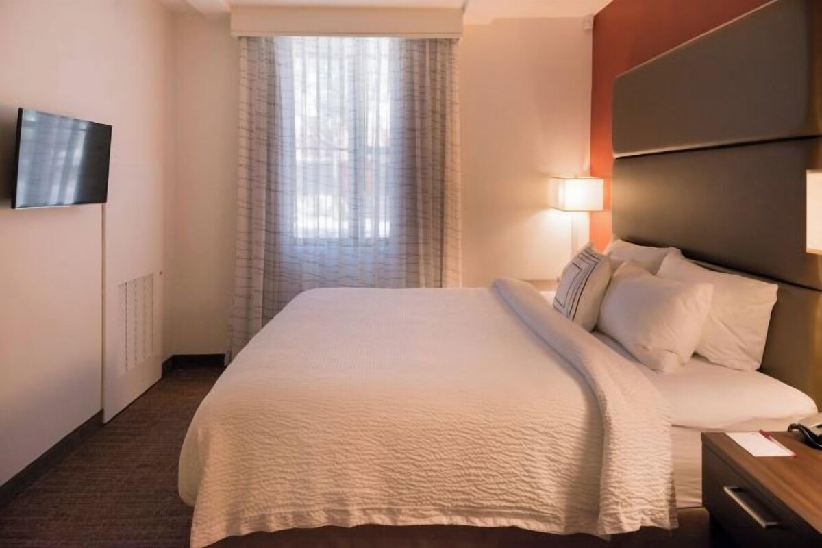 Habitación de hotel con una cama grande, un televisor de pantalla plana montado en la pared, una lámpara de noche y una ventana con cortinas translúcidas.