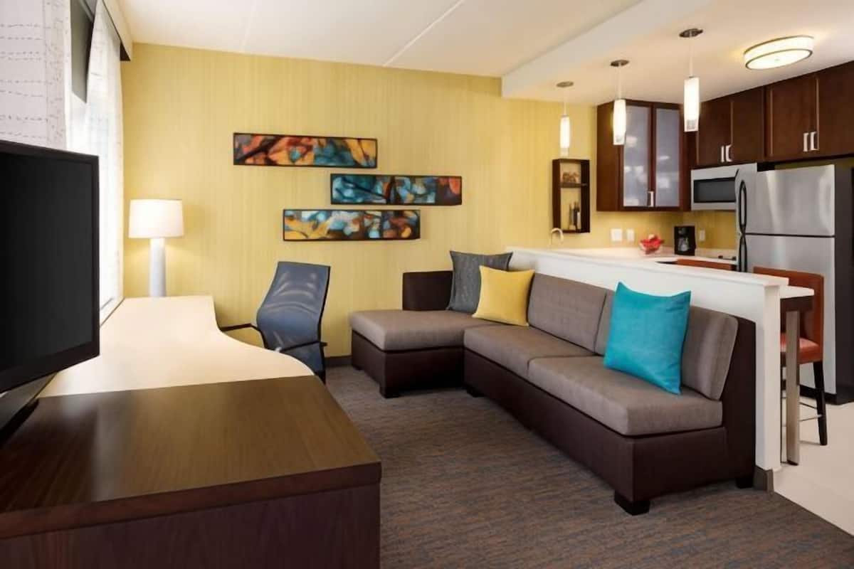 A hotel room with a sofa, a TV, a desk, a kitchen area, and artwork on the wall.