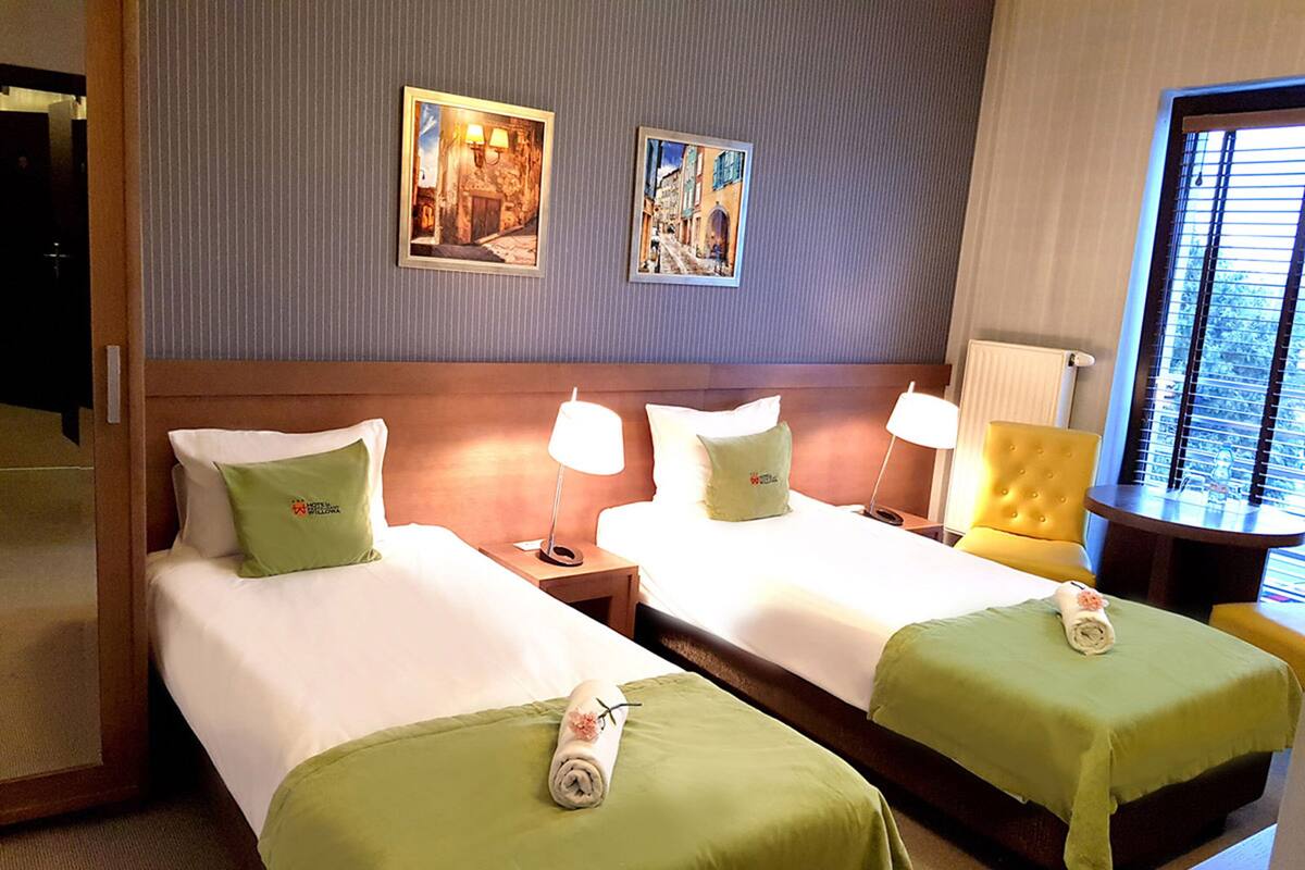 Una habitación de hotel con dos camas, cada una con una colcha verde y una toalla enrollada. Hay dos lámparas de pared en cada mesita de noche, y un cuadro sobre las camas.