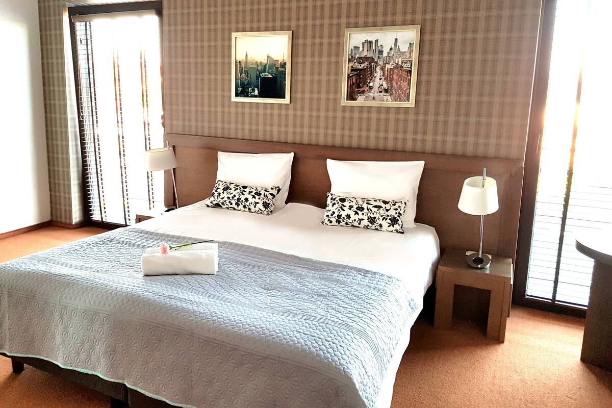Habitación de hotel con una cama grande, dos almohadas, una mesita de noche con lámpara y dos cuadros enmarcados en la pared.