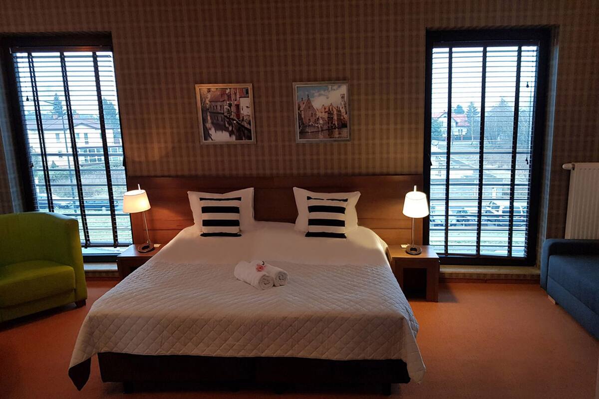 Habitación de hotel con una cama grande, dos lámparas de noche, un sillón verde y dos ventanas con vistas a la ciudad.