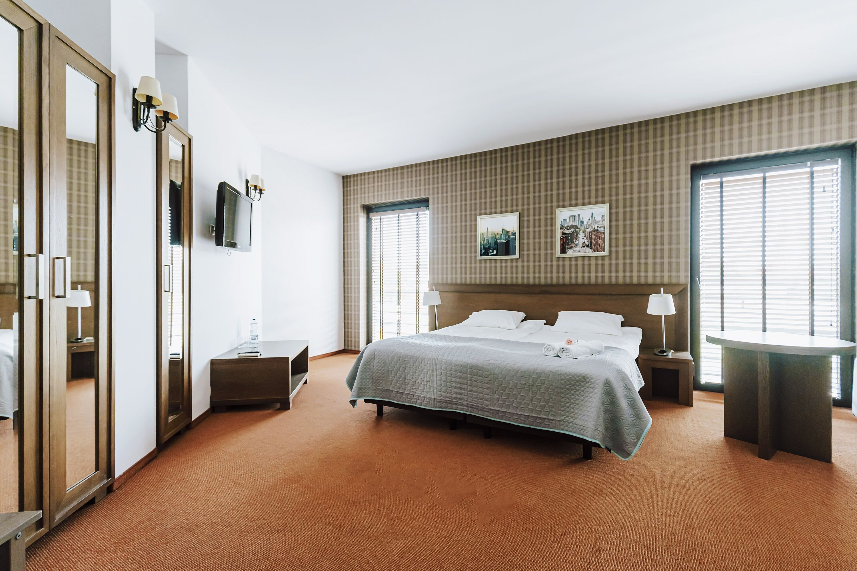 A hotel room with a large bed, a bedside table, a mirror, a TV, and a window with blinds.