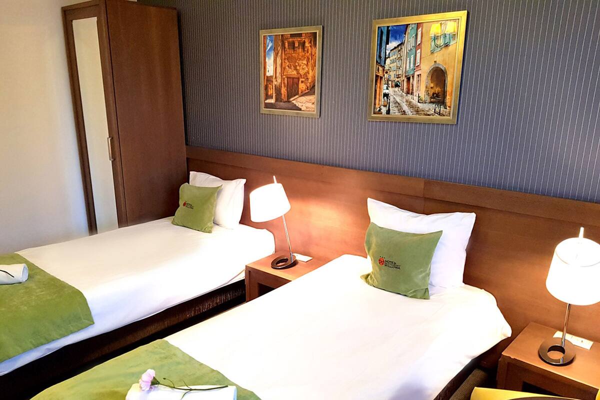 Una habitación de hotel con dos camas, cada una con un almohadón verde y uno blanco, un cabecero de madera y una mesita de noche con una lámpara.