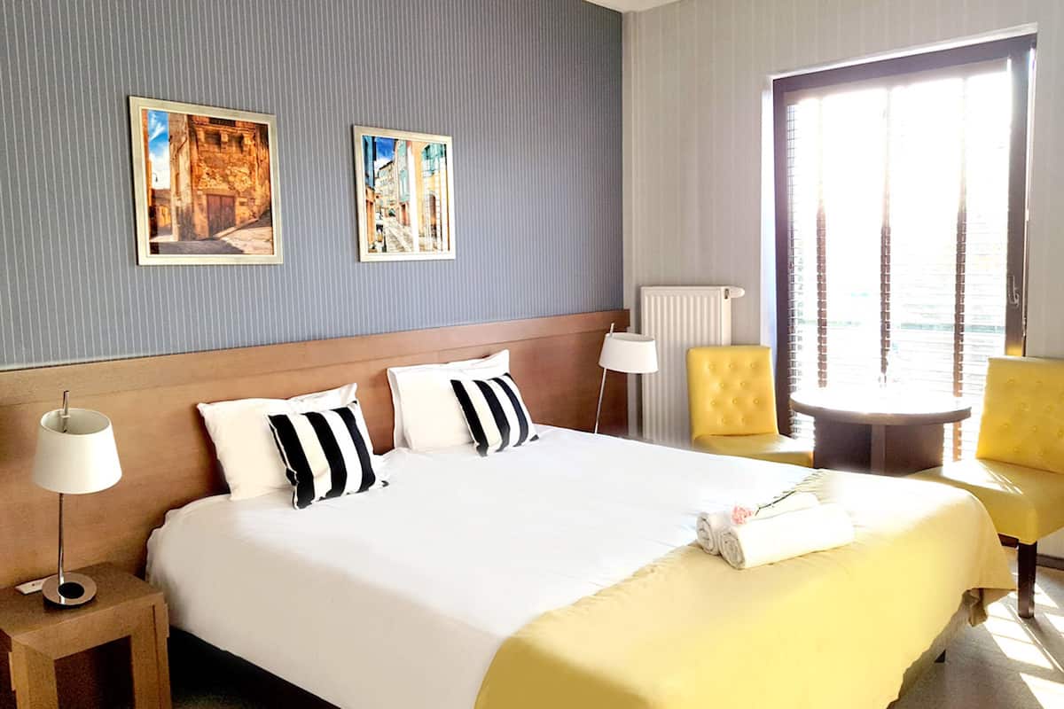 Habitación de hotel con una cama grande, dos mesitas de noche, una silla y una ventana con persianas.