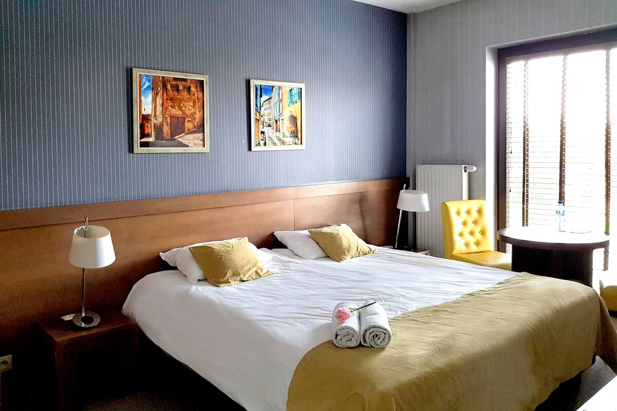 Habitación de hotel con una cama grande, una mesita de noche, una lámpara, una silla y dos cuadros en la pared.