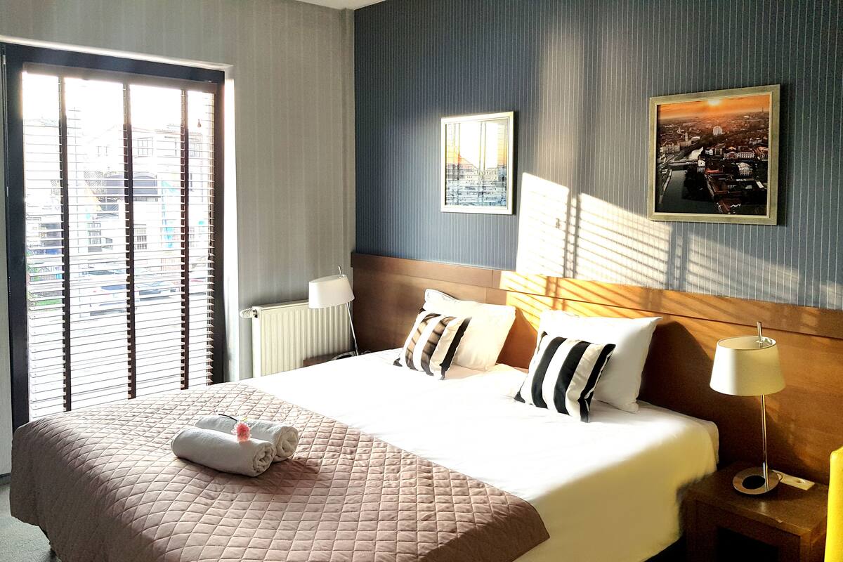 Una habitación de hotel con una cama grande, una mesita de noche, una lámpara y una ventana con persianas.