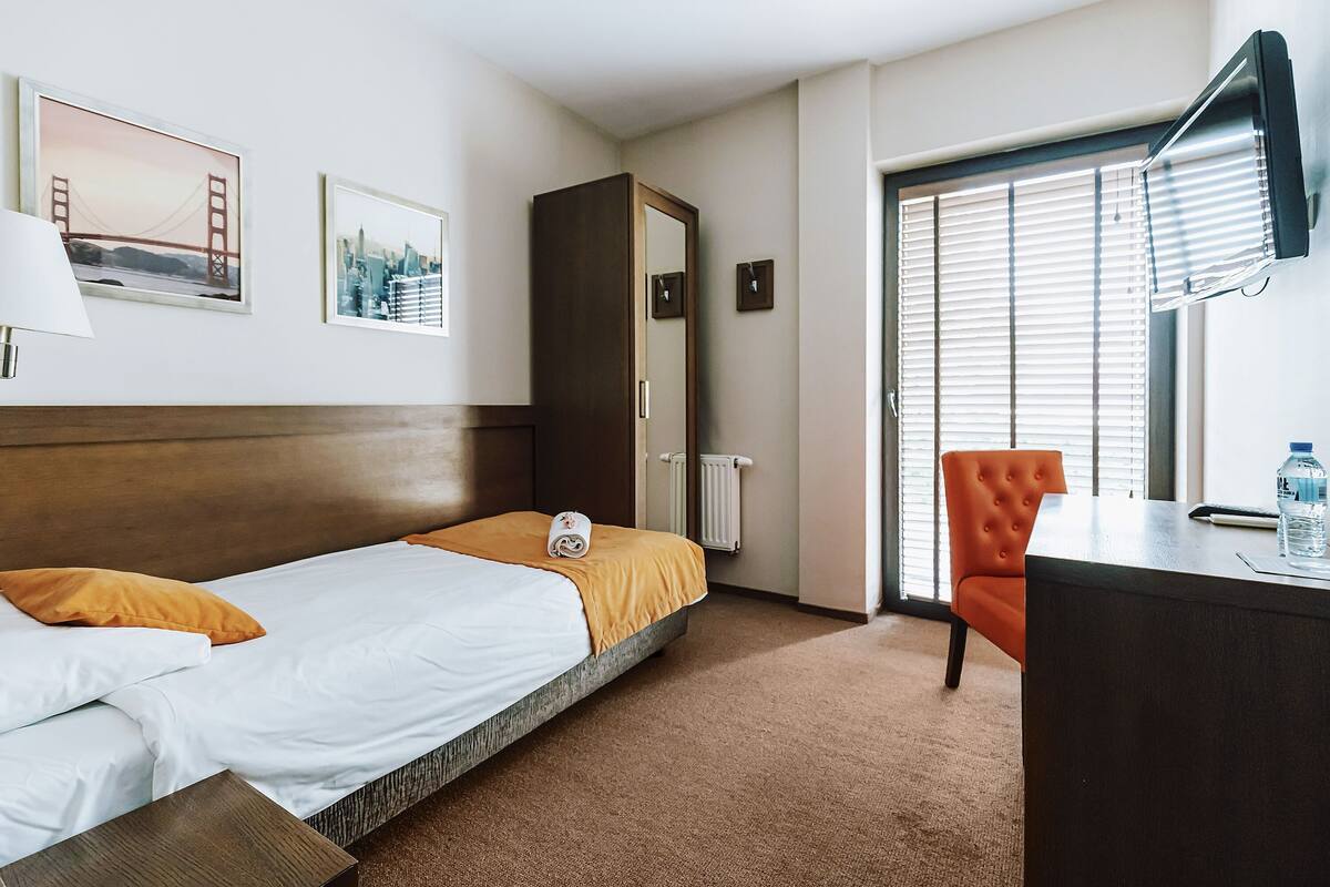 Habitación de hotel con cama, escritorio, silla, radiador y ventana con persianas.