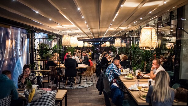 Un restaurante con mesas y sillas de madera, plantas en macetas y lámparas colgantes.