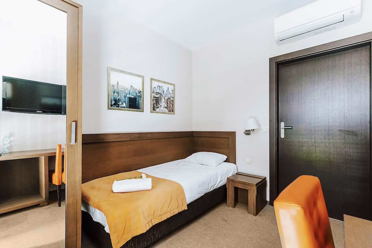 Habitación de hotel con una cama individual, un escritorio con silla, televisión, dos cuadros en la pared y una puerta.