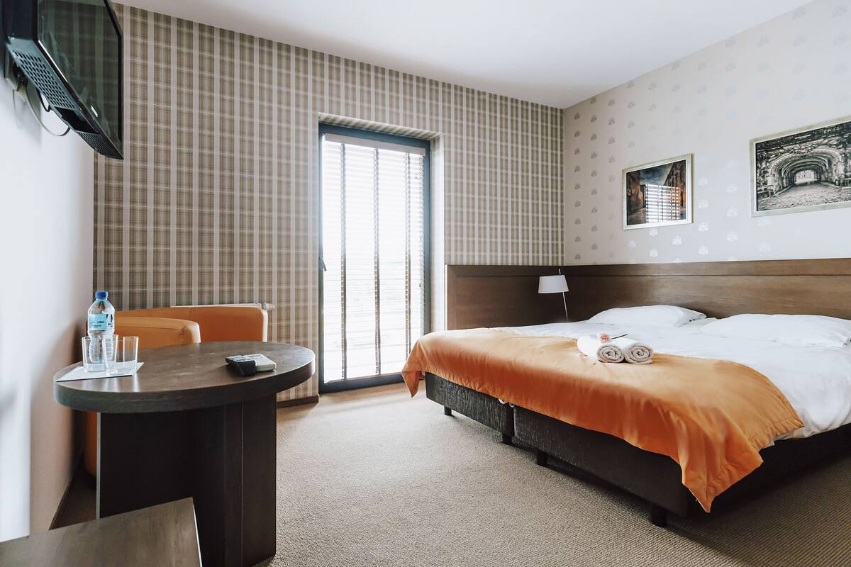 Habitación de hotel con cama, escritorio, silla, televisor y ventana con persianas.
