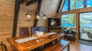Een houten interieur met een eettafel, stoelen, een televisie en een groot raam met uitzicht op een meer.
