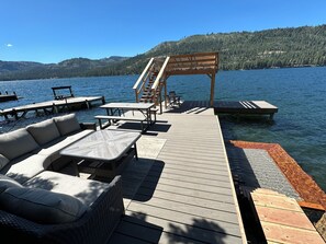 A wooden deck with outdoor seating, including a sofa and tables, overlooking a lake with docks and a forested hillside in the background. - Lakefront Cabin with private pier on Donner Lake (Truckee)