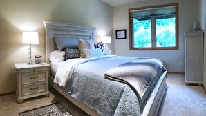 Un dormitorio con cama, mesitas de noche, lámpara, cómoda y ventana con vista a árboles.