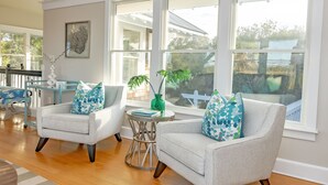 A spacious living room with two light grey armchairs, a small round table, and a large window offering a view of the outdoors. - Get ready for an unforgettable marsh view at 3 Little Birds on Bald Head Island! (Bald Head Island)