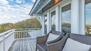 A wooden deck with wicker furniture and large windows overlooking a natural landscape. - Get ready for an unforgettable marsh view at 3 Little Birds on Bald Head Island! (Bald Head Island)