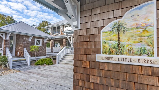 A wooden deck leading to a house with a sign reading "THREE LITTLE BIRDS." - Get ready for an unforgettable marsh view at 3 Little Birds on Bald Head Island! (Bald Head Island)