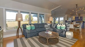 A spacious living room with a sectional sofa, a round coffee table, and a view of a field through large windows. - Get ready for an unforgettable marsh view at 3 Little Birds on Bald Head Island! (Bald Head Island)