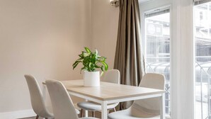 A modern dining area with a light-colored table and four chairs, a potted plant, and a view of the city through large windows.