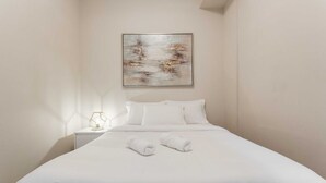 A neatly made bed with white linens and two rolled towels, a bedside lamp, and a wall-mounted abstract painting.