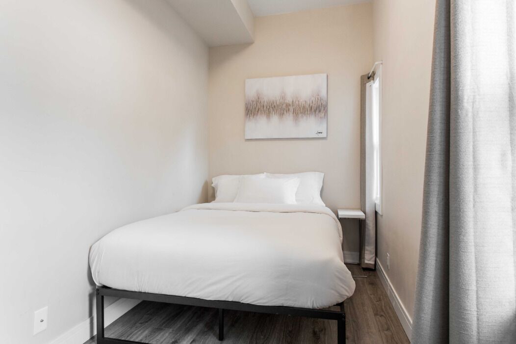 Un dormitorio pequeño y ordenado con cama, mesita de noche, una obra de arte en la pared y ventana con cortinas.