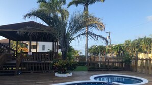 Zona de piscina con cubierta de madera, espacio de descanso techado y jardín cercado con palmeras.