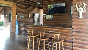 Interior de una cabaña de troncos de madera con una zona de bar que incluye taburetes de madera y un televisor de pantalla plana montado en la pared.