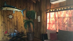 Un interior de madera con techo de madera, una mesa de vidrio con objetos decorativos, un sofá, una planta en maceta y un aire acondicionado instalado en la pared.