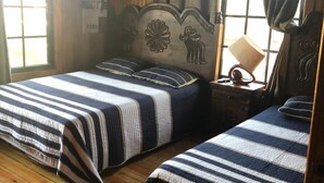 Habitación con dos camas, suelo de madera, cabecera con adornos tallados, mesita de noche con lámpara y ventana con cortinas.