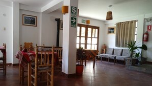 Amplia habitación con zona de comedor, sillas de madera y una mesa cubierta con mantel rojo. Cuenta con un conjunto de sofá y varias plantas en macetas. Un letrero indica una salida.