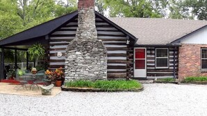 Une cabane rustique en rondins de bois, dotée d’une cheminée en pierre, d’un patio couvert avec des meubles d’extérieur et d’une allée en gravier.