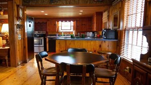 L’intérieur d’un chalet en bois comprend une table à manger ronde et des chaises, une cuisine avec des meubles en bois et une grande fenêtre équipée de stores.
