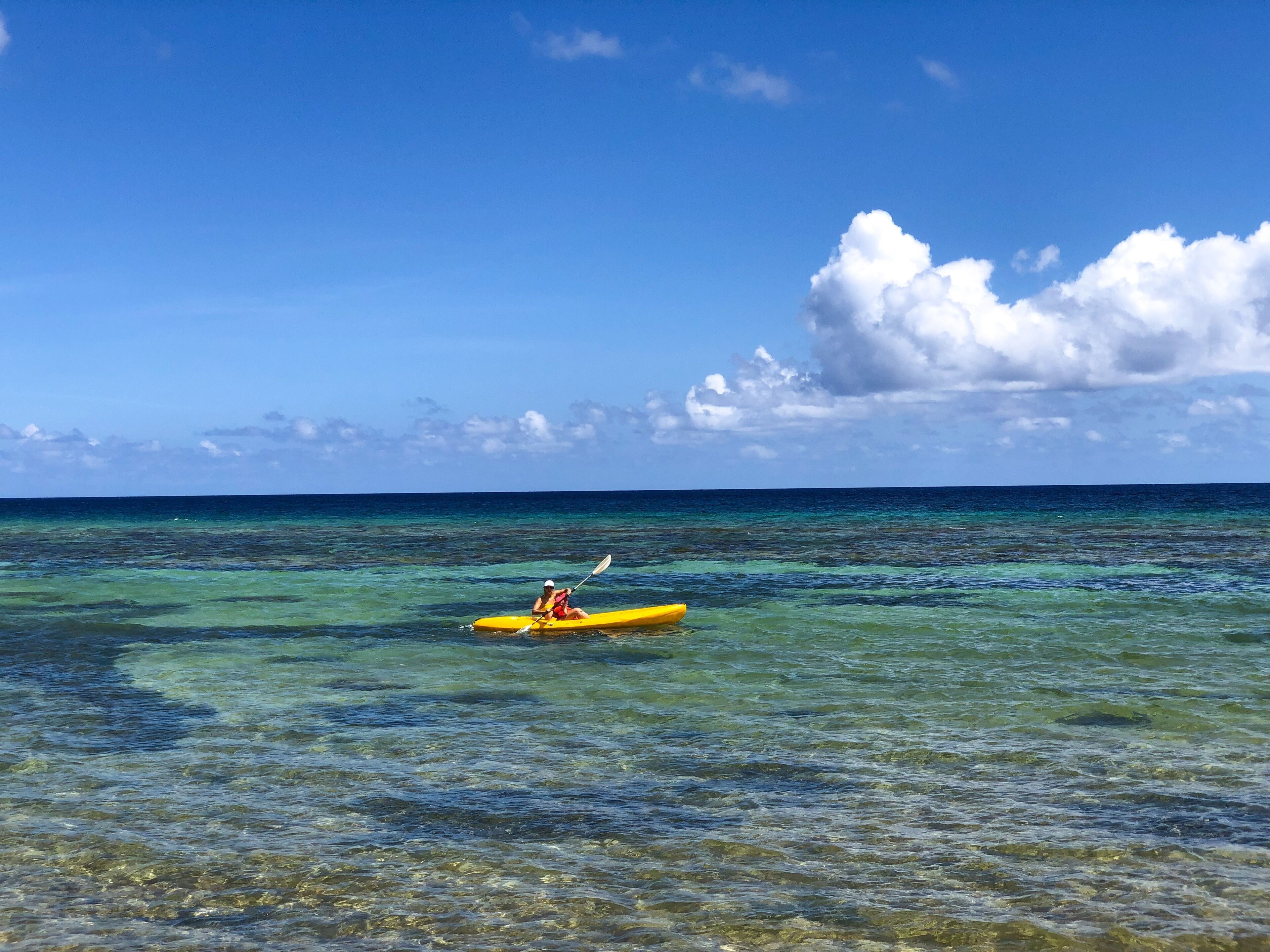 Un kayak sur une eau turquoise et limpide, avec une personne qui rame, sous un ciel bleu vif parsemé de quelques nuages.